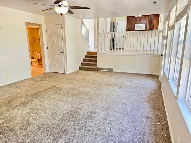 Carpeted living area featuring a ceiling fan with integrated lighting, multiple windows with louvered shutters, and a staircase with a white baluster railing