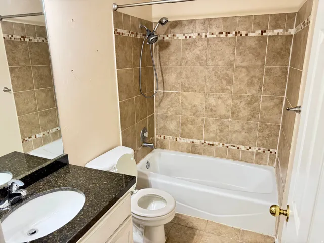 Bathroom featuring a built-in bathtub with a shower combination, neutral-toned wall tiles with a decorative mosaic accent band, a vanity with a dark speckled countertop, and a white integrated sink