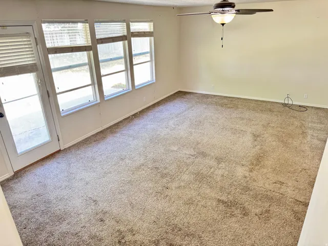 Spacious room featuring neutral carpeting, a ceiling fan with integrated lighting, multiple windows, and a glass-paneled door