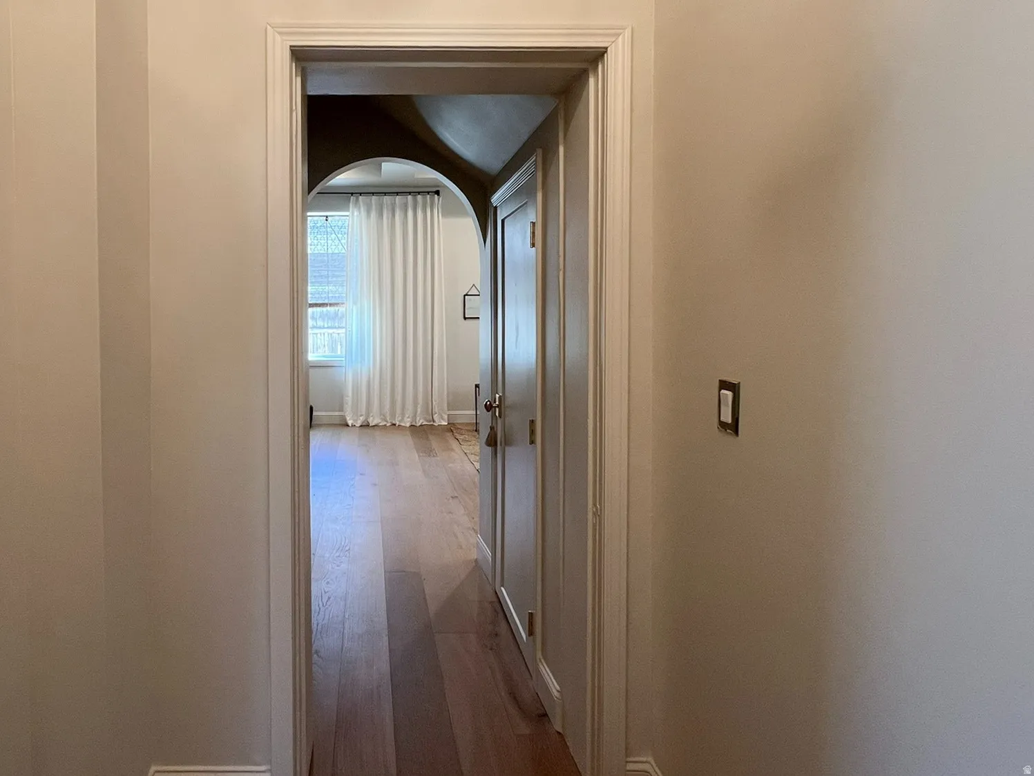 Hallway entry to a room featuring an arched doorway, wood-finish flooring, and a window with drapery