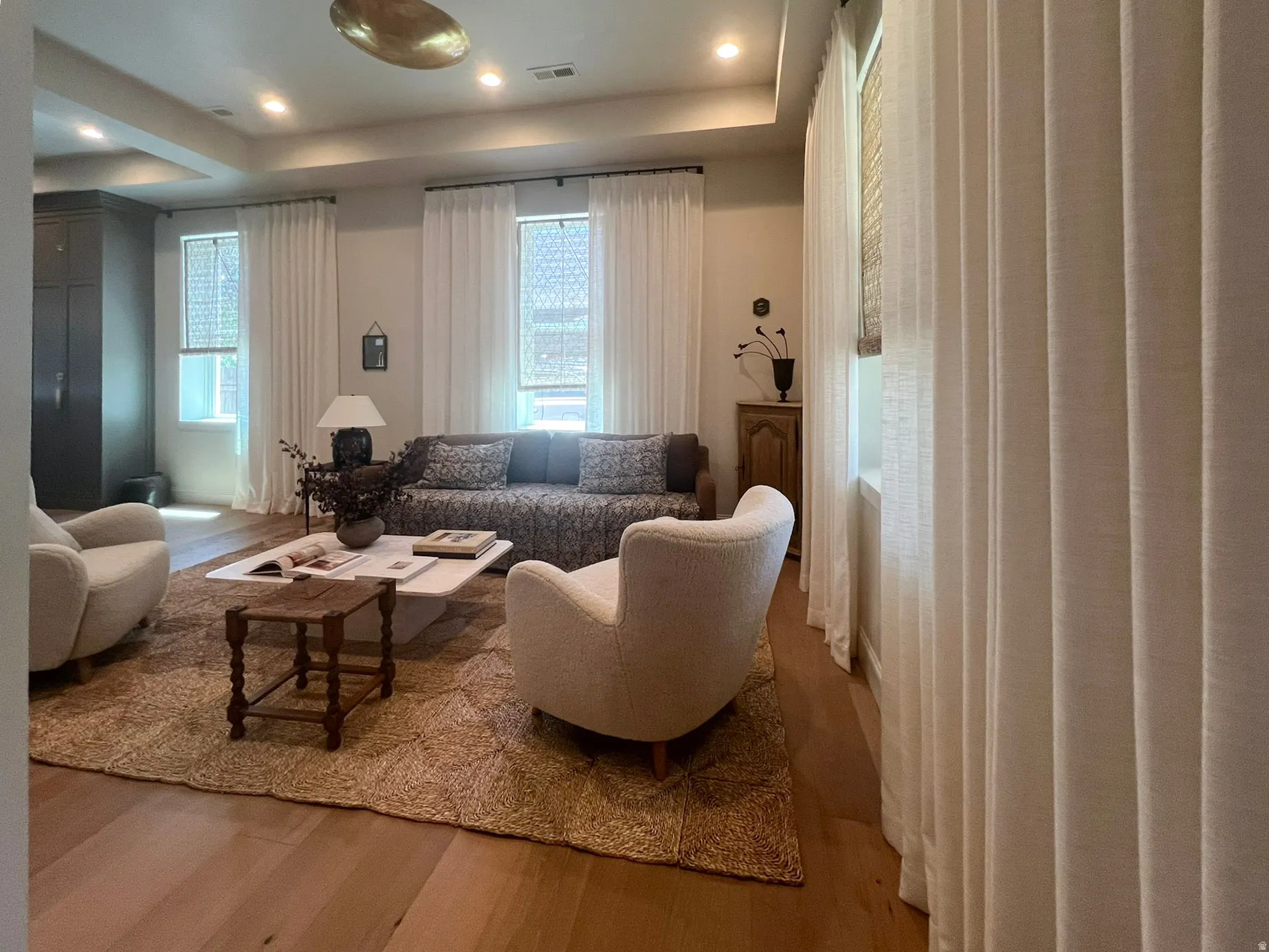 Spacious living area featuring wood-finish flooring, a tray ceiling with recessed lighting, and large windows with privacy blinds and drapes