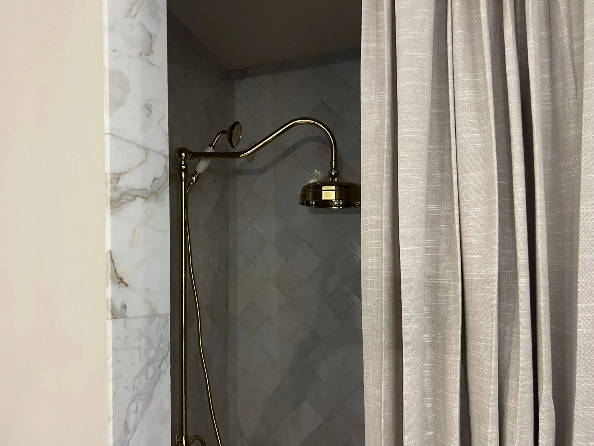 Built-in shower featuring a brass-finish rainfall showerhead and handheld sprayer, surrounded by large-format stone-finish wall tiles and diagonal-set gray shower tiles