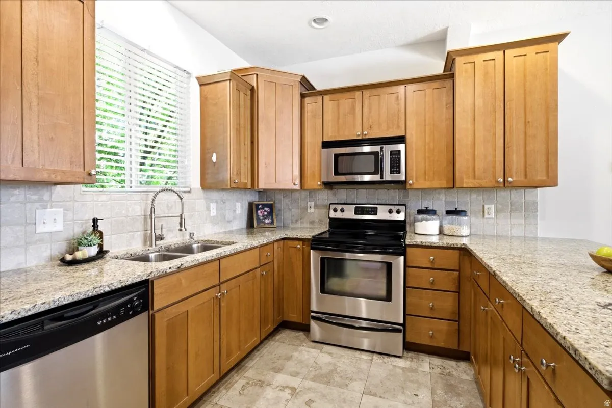 Kitchen featuring wood cabinetry, stainless steel appliances, granite countertops, a double basin sink, and a tile backsplash