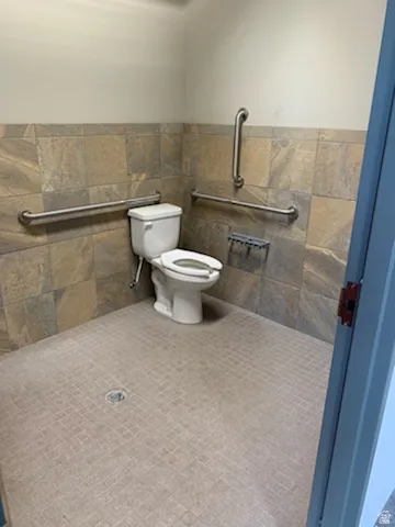 Tiled restroom featuring a white toilet, full wall tiling with a neutral palette, and stainless steel grab bars