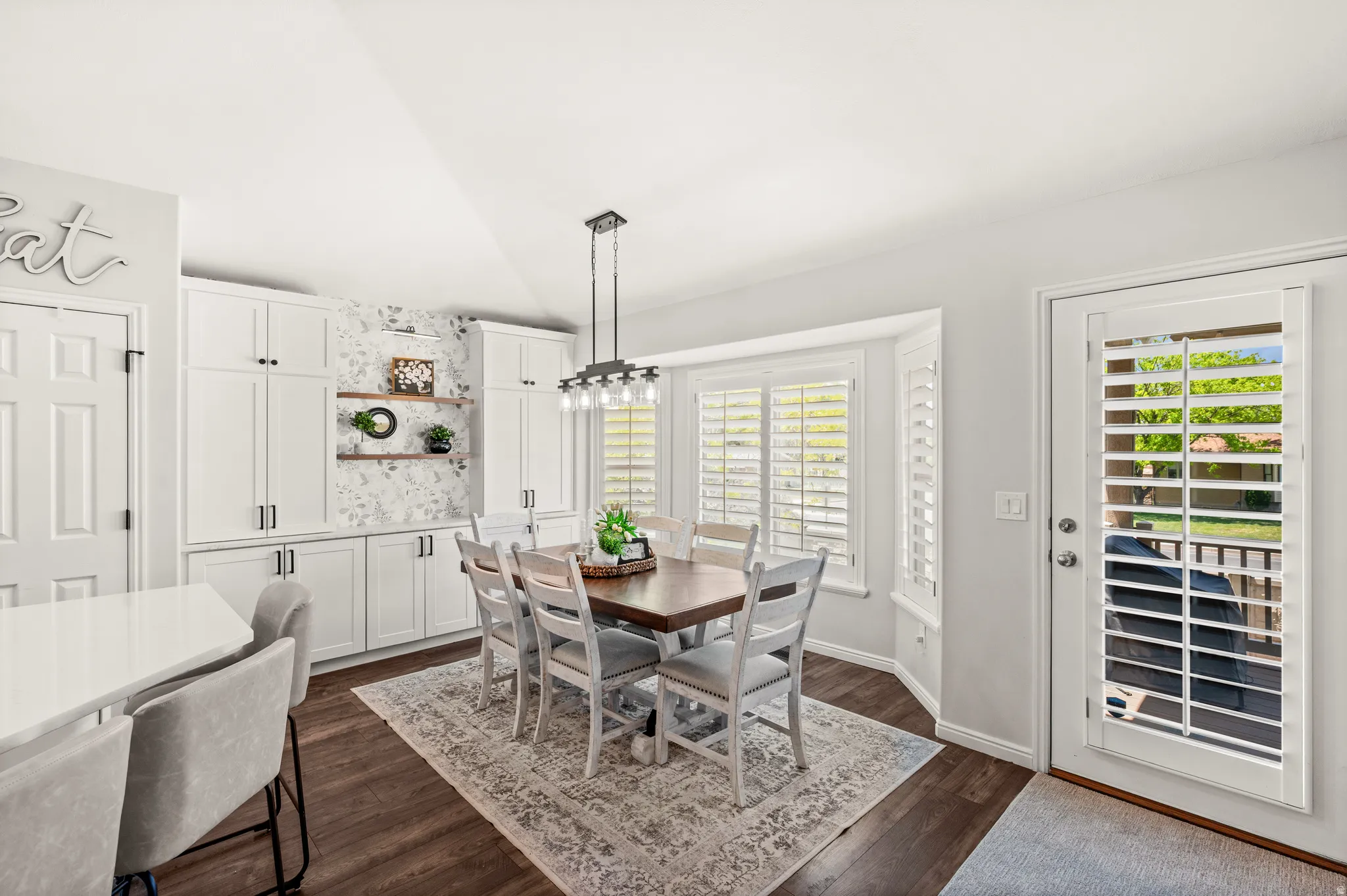 Modern dining space featuring white shaker cabinetry with dark hardware, recessed shelving with a patterned wall accent, and a bay window with plantation shutters