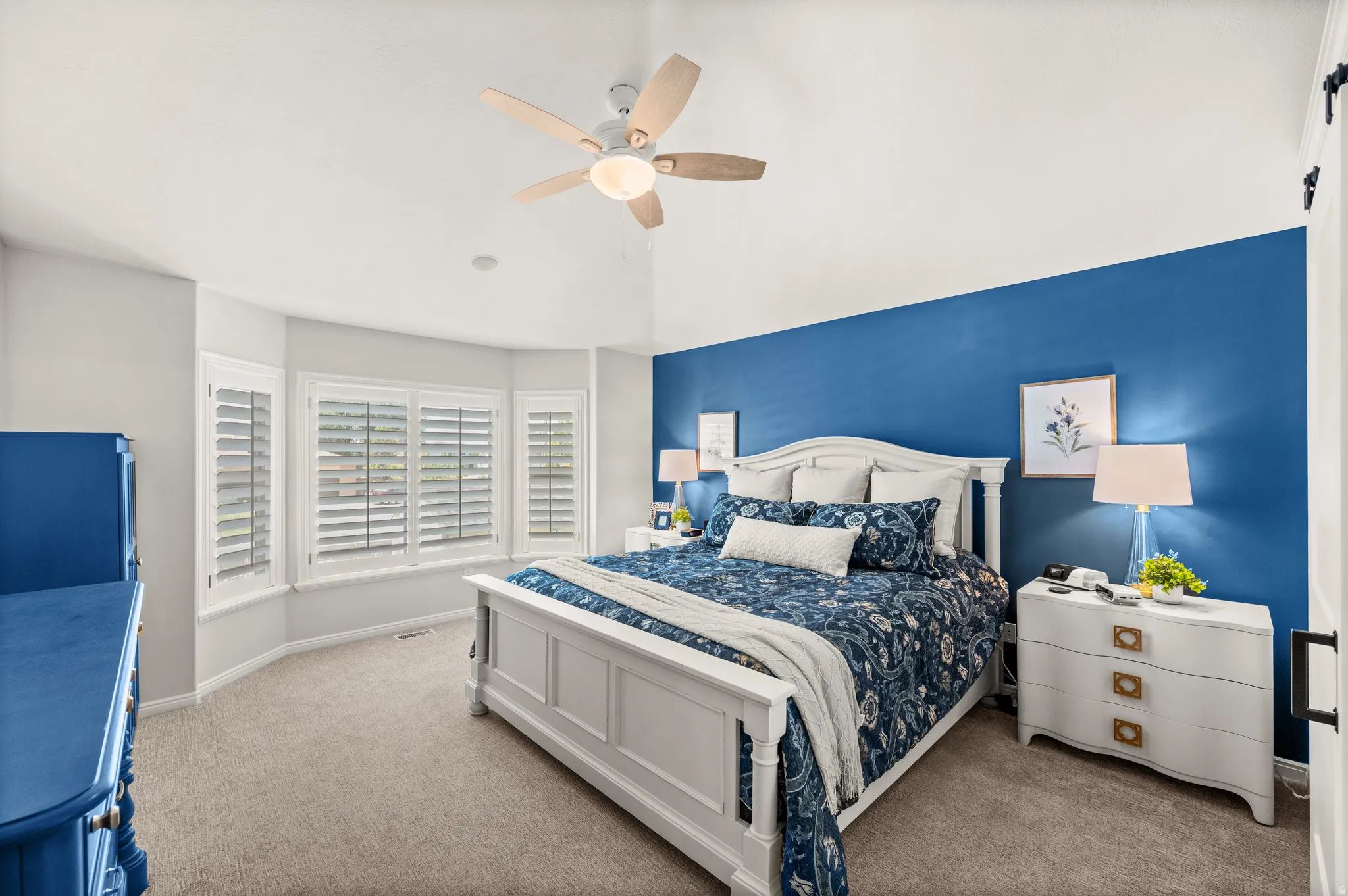 Vaulted ceiling bedroom featuring a bay window with plantation shutters, ceiling fan, and carpet flooring