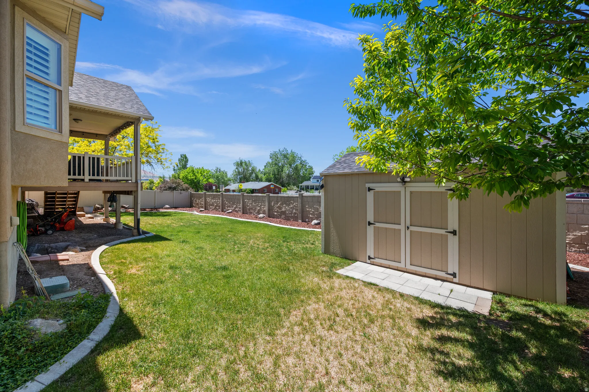 Expansive backyard featuring a manicured lawn, an elevated covered deck with white railings, a concrete patio area, and a detached storage shed with double doors
