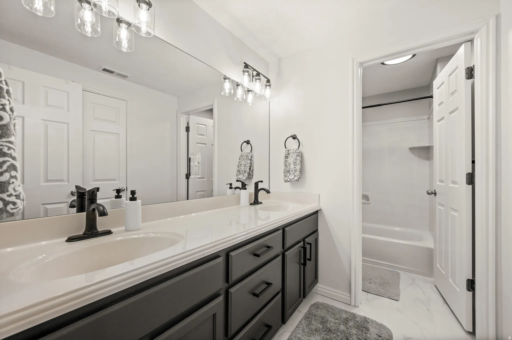Bathroom featuring a dual vanity with a light-toned countertop, matte black fixtures, dark cabinetry, and an expansive wall mirror