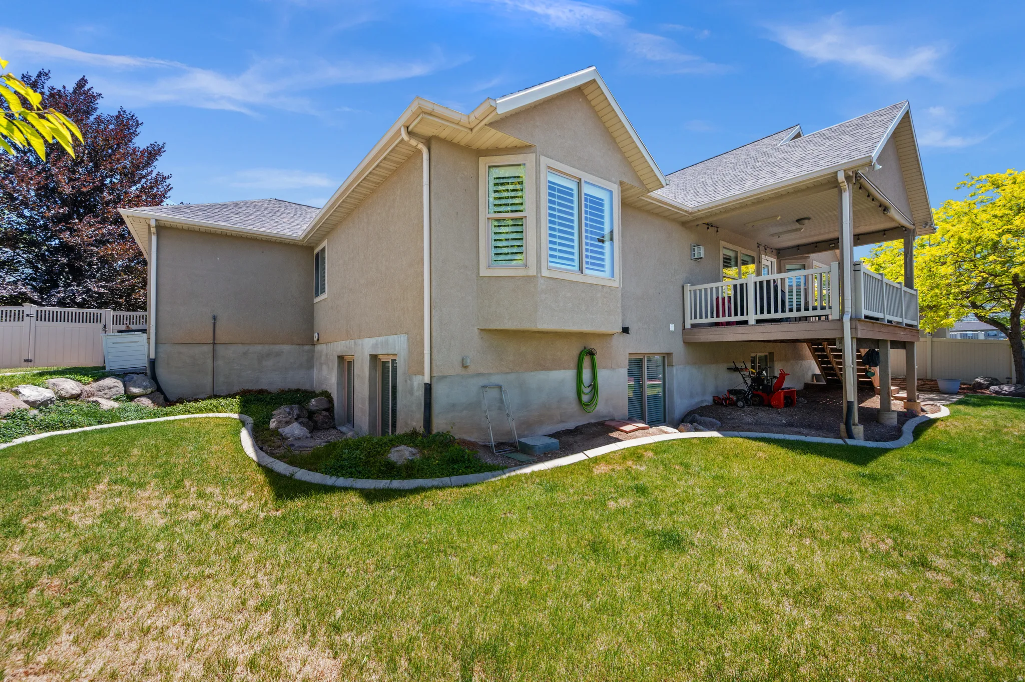 Rear exterior featuring a covered deck with white railings, a lower-level concrete patio, and a landscaped yard with a rock border