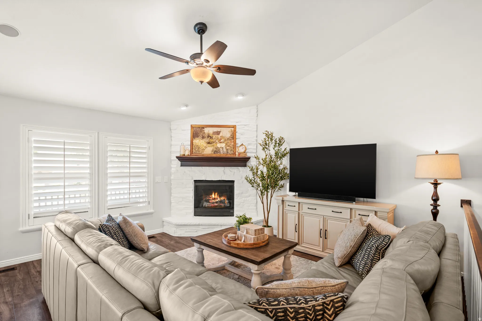 Living room featuring a corner fireplace with a white-painted brick surround and dark wood mantel, wood-finish flooring, white walls, and a ceiling fan with integrated lighting