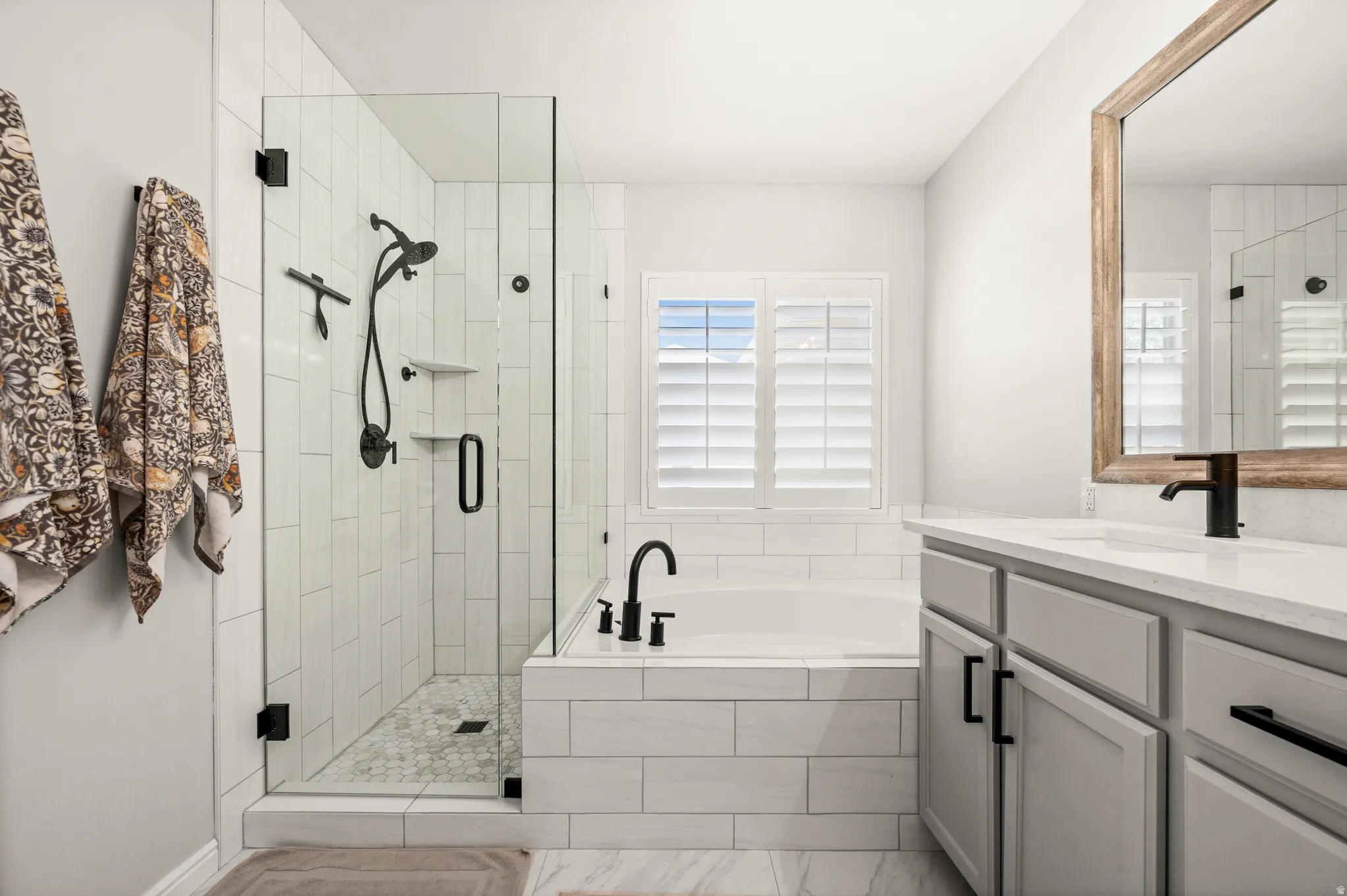 Bathroom featuring a walk-in shower with vertical subway tile, a freestanding soaking tub with tile surround, a single vanity with gray cabinetry, a light-toned countertop, and a framed mirror
