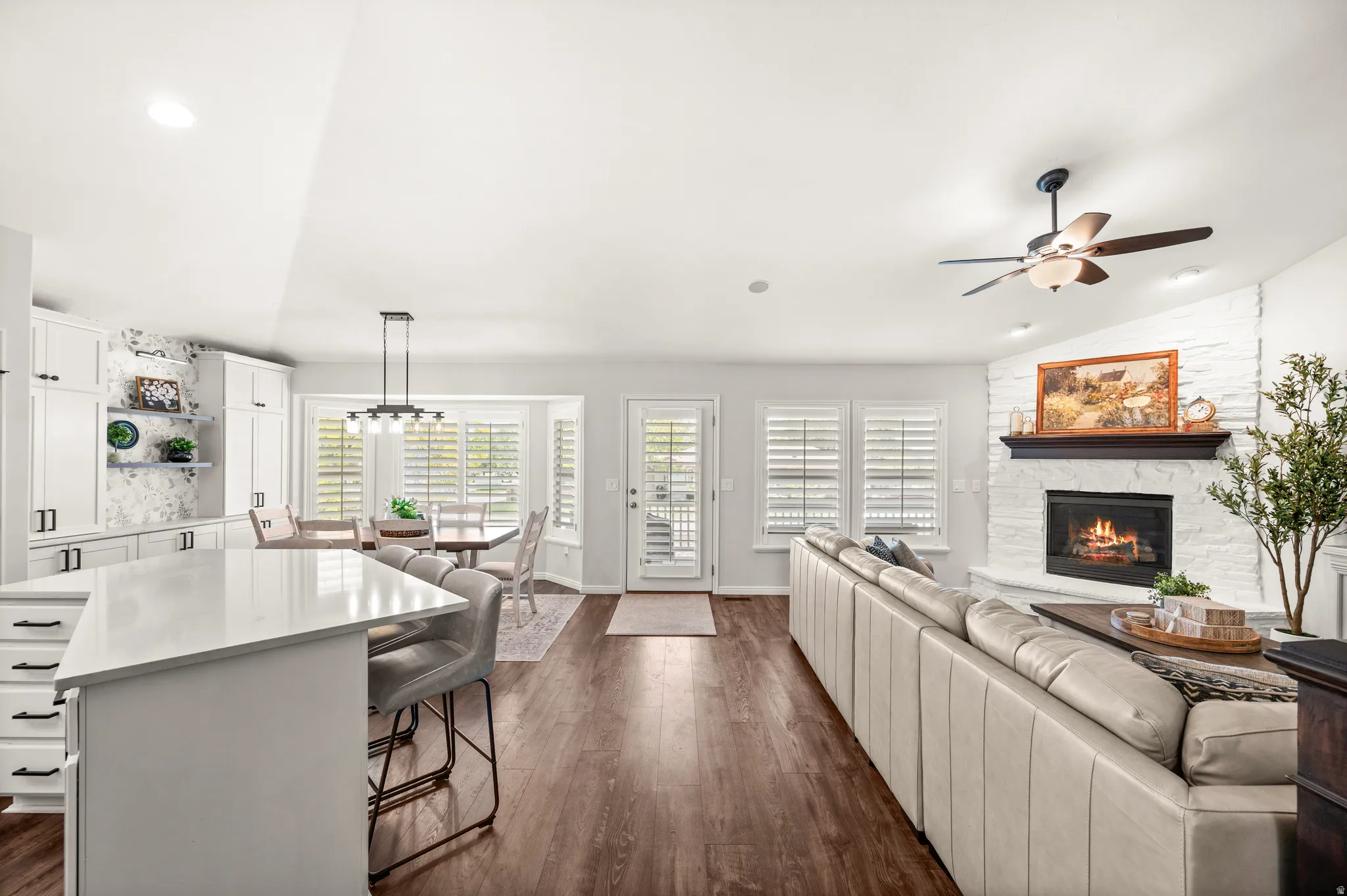 Open concept living area featuring wood-finish flooring, a white brick fireplace with dark wood mantle, white kitchen cabinetry with dark hardware, a kitchen island with seating, and a bay window in the dining area
