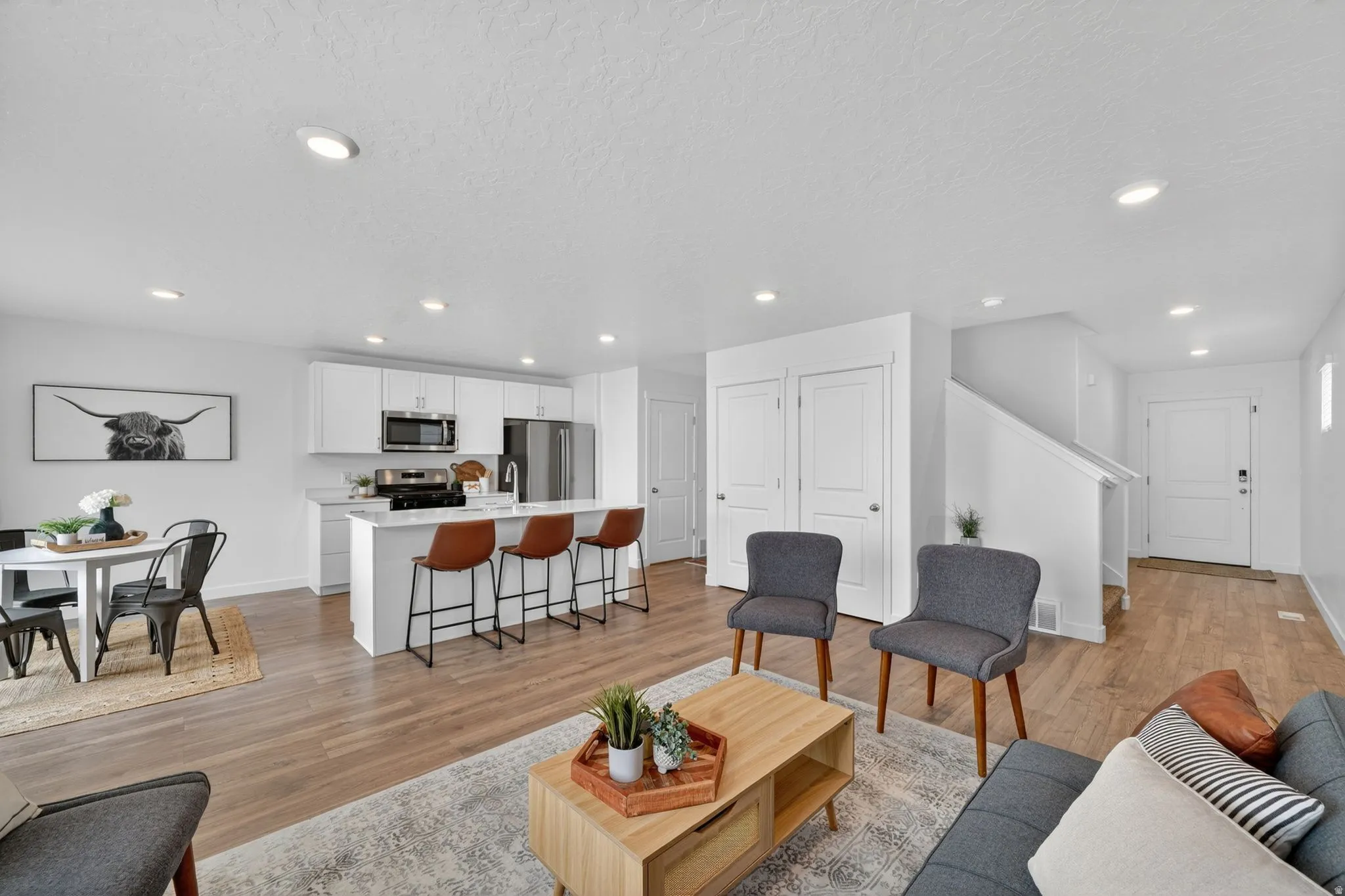 Open-concept living space with wood-finish flooring, recessed lighting, and a white kitchen featuring a large island with seating