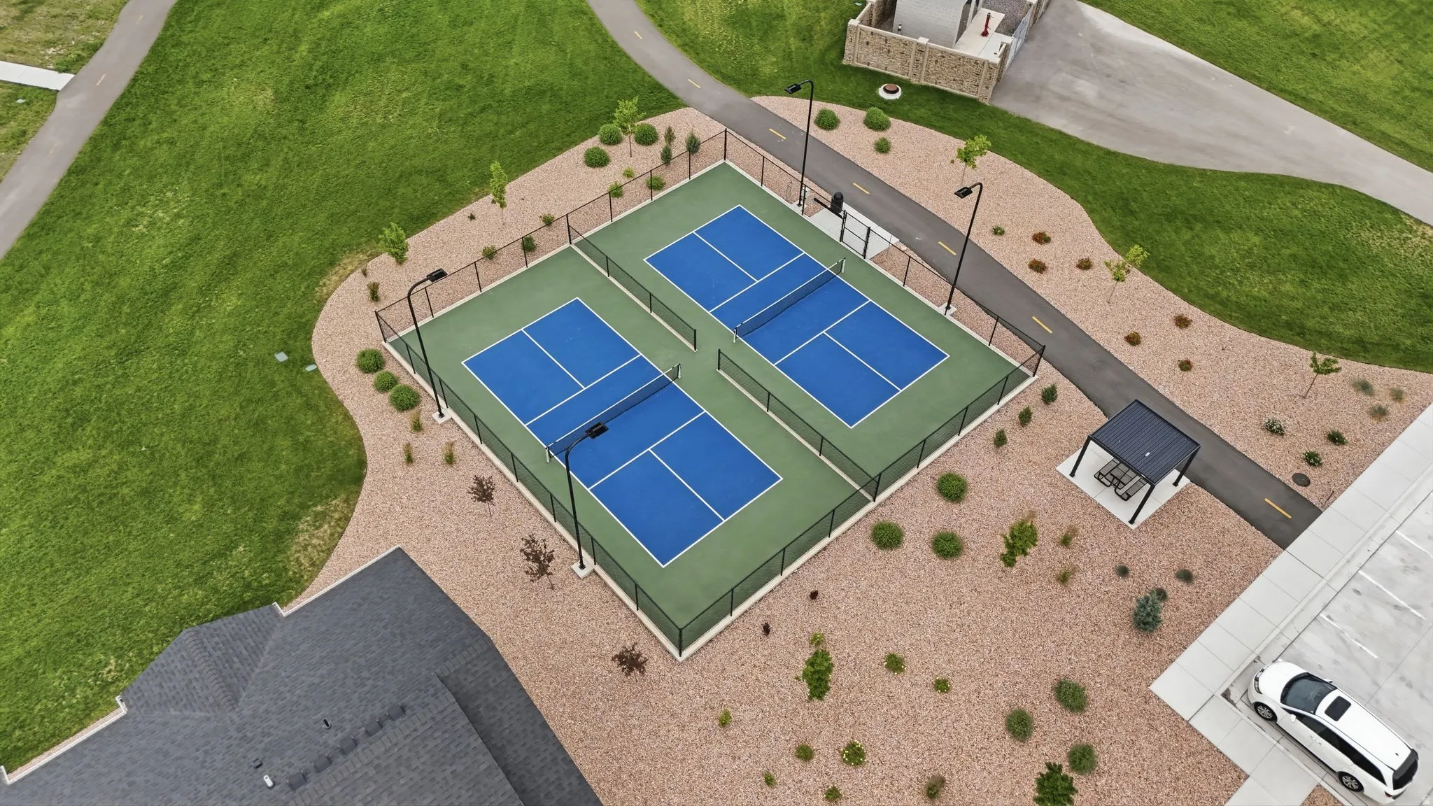 Two fenced blue and green pickleball courts with overhead lighting, bordered by landscaped gravel and mature grass