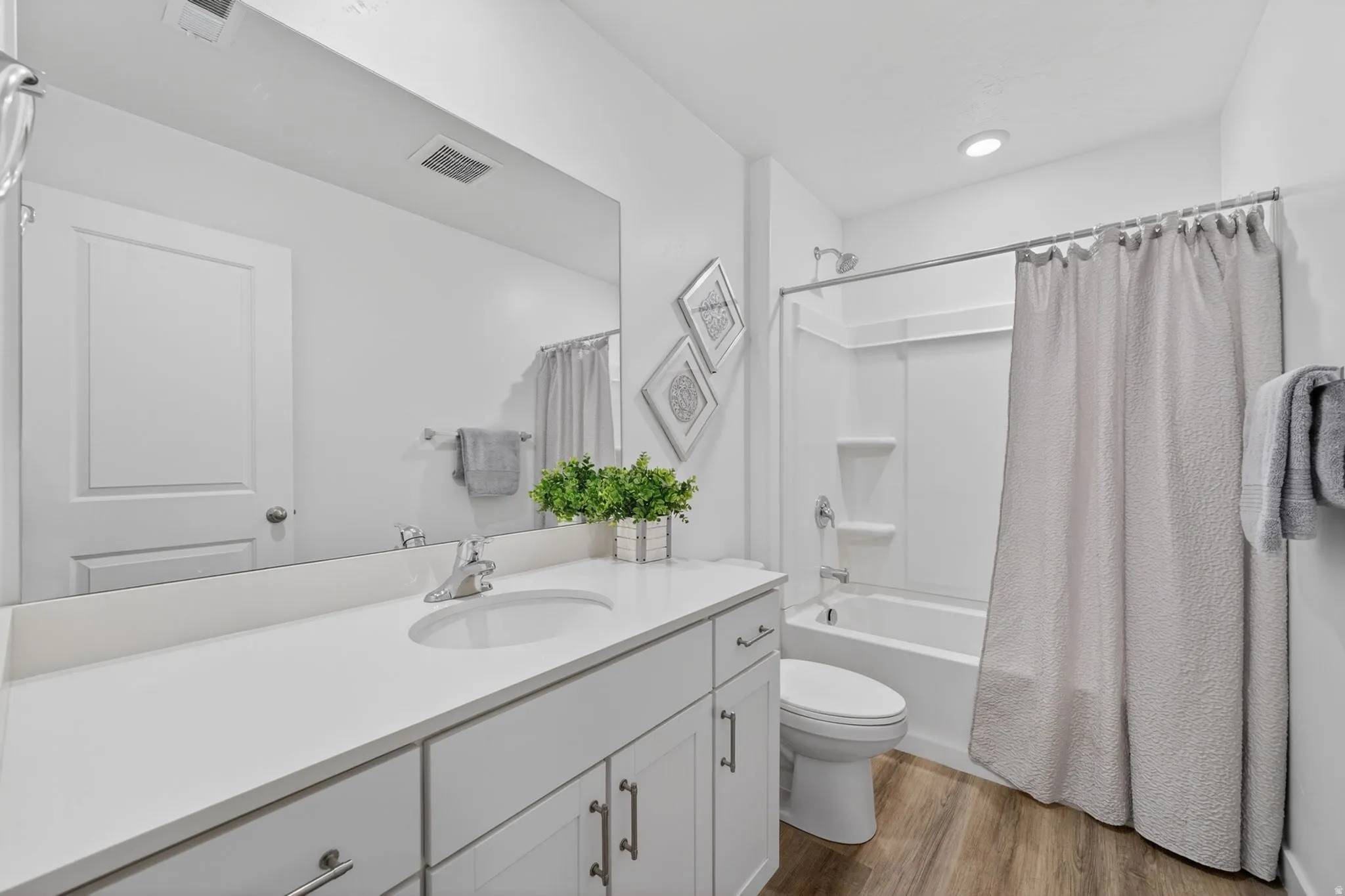 Contemporary bathroom featuring a white vanity with integrated sink, a large wall-mounted mirror, a tub/shower combination with a white surround, and wood-finish flooring