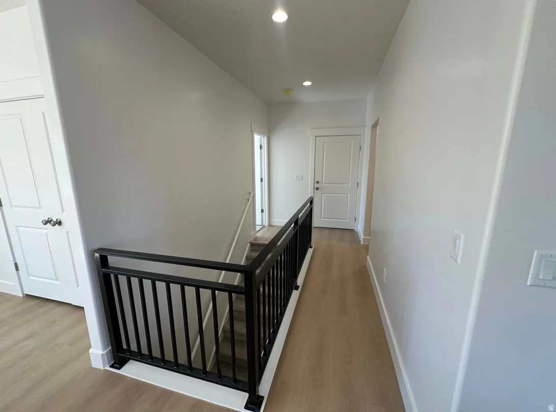 Upper-level hallway featuring light wood-finish flooring, recessed lighting, white walls, and a contemporary black metal staircase railing