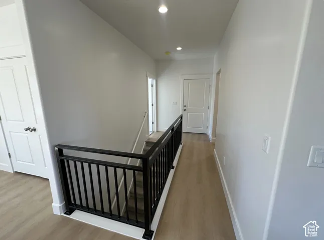 Upper-level hallway featuring light wood-finish flooring, recessed lighting, white walls, and a contemporary black metal staircase railing