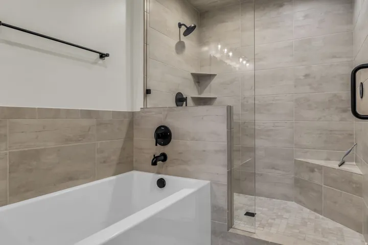 Modern bathroom featuring a soaking tub, glass-enclosed walk-in shower, dark-finish fixtures, light-toned tile surrounds, and integrated shower seating
