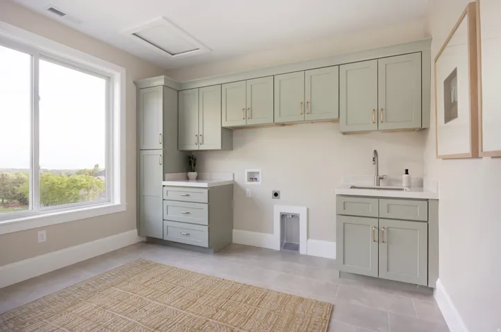 Laundry area featuring extensive sage green cabinetry with gold-finish hardware, a large window, a built-in sink with a gooseneck faucet, light-toned countertops, and light gray tile flooring