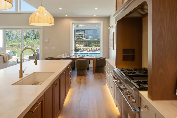 Gourmet kitchen featuring an integrated gas range with a custom wood-paneled hood, a large center island with an undermount sink and brass-finish faucet, under-cabinet lighting, wood-finish flooring, and a dining area with direct access to an outdoor pool