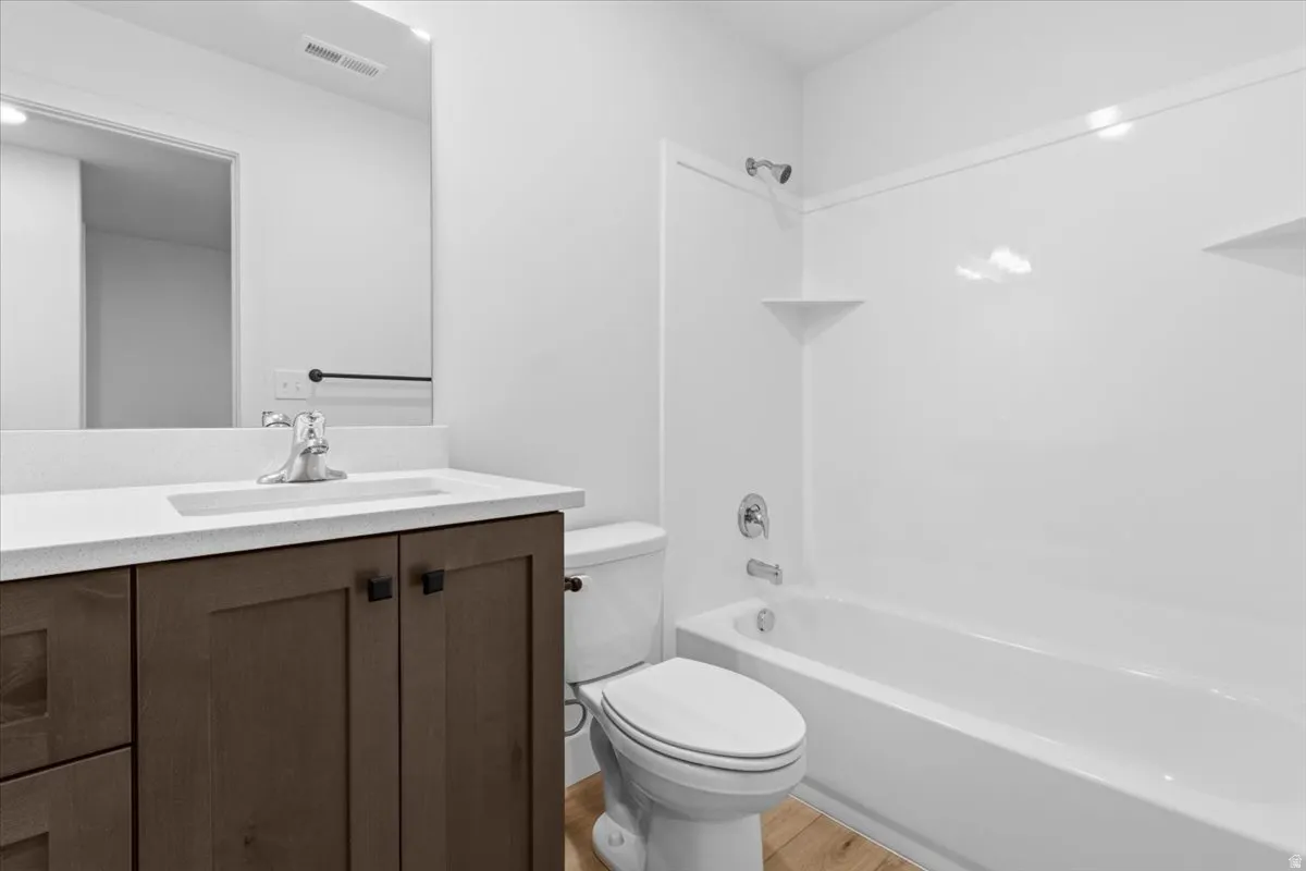 Bathroom featuring a wood-finish vanity with a white countertop and integrated sink, a chrome faucet, a large wall-mounted mirror, a white bathtub with an integrated shower, and wood-finish flooring