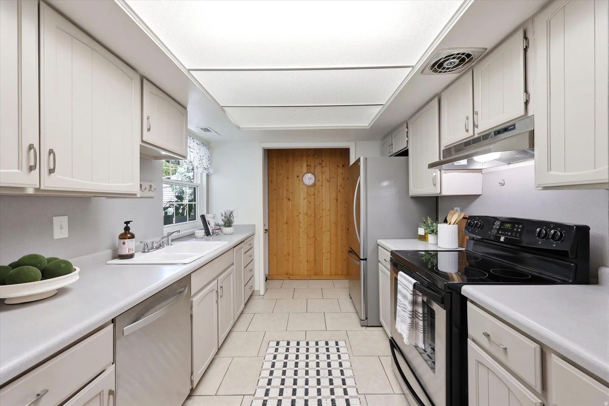 Galley kitchen featuring light cabinetry with brushed nickel hardware, a stainless-steel dishwasher, a black electric range with an overhead microwave, a stainless-steel refrigerator, and light tile flooring