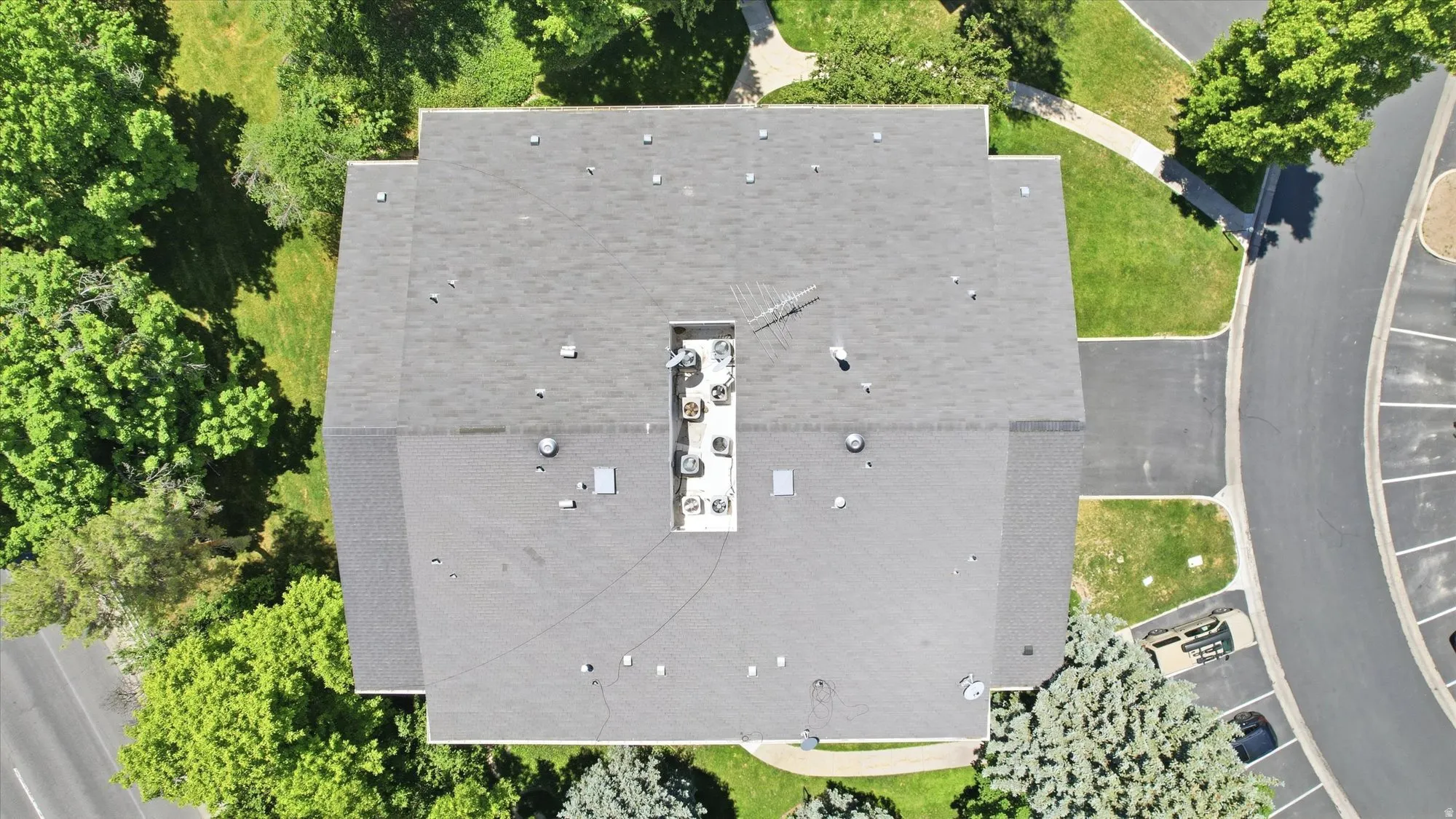 Rectangular building featuring a dark shingle roof, roof-mounted mechanical units, and surrounding green spaces with mature trees