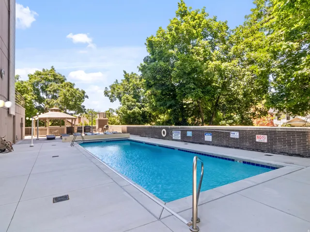 Outdoor swimming pool with concrete decking, a brick privacy wall, surrounding mature trees, and an adjacent gazebo