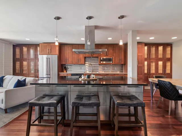 Spacious kitchen featuring an expansive island with a dark countertop, stainless steel appliances, a geometric tile backsplash, and extensive wood cabinetry with glass-front uppers