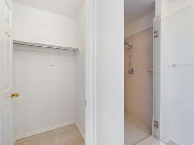 Recessed closet space with a clothing rod and light-toned tile flooring, complemented by a nearby tiled shower enclosure with a handheld showerhead and built-in shelf
