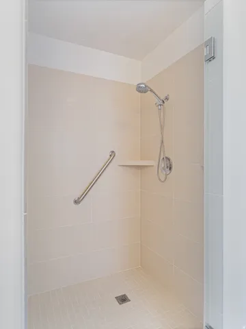 Walk-in shower with neutral wall tiling, a handheld showerhead, corner shelf, and grab bar
