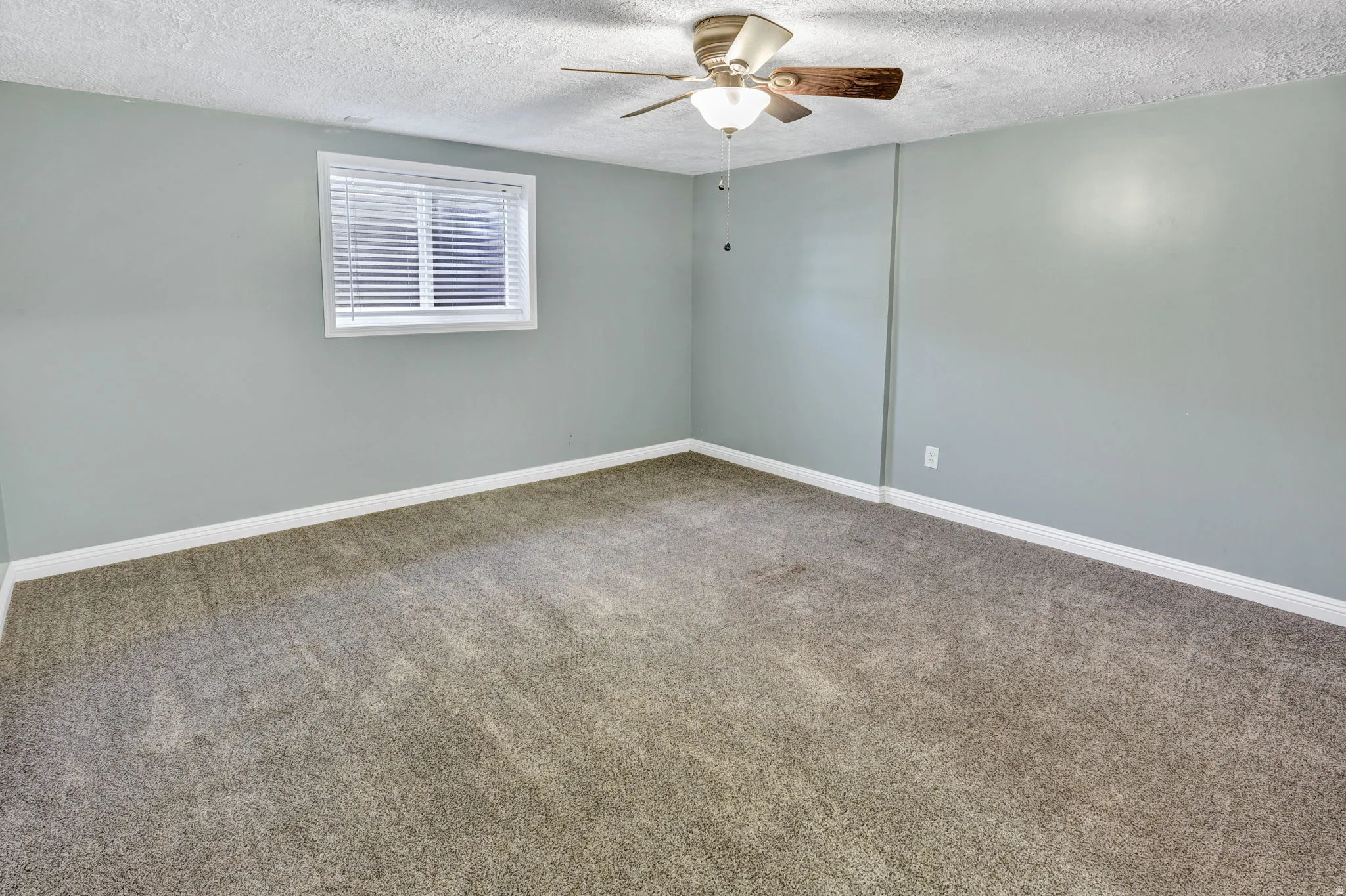 Carpeted interior room featuring light gray walls, white trim, a single window with blinds, and a ceiling fan with light fixture