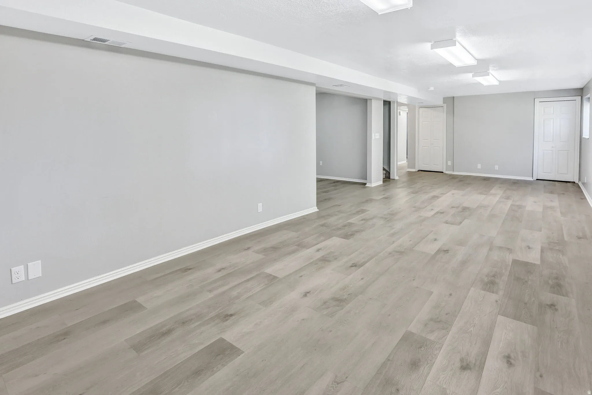 Spacious room featuring light wood-finish flooring, neutral gray walls, white baseboards, and overhead fluorescent lighting