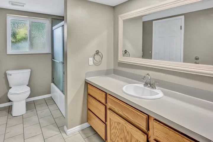 Bathroom featuring a wood-finish vanity with an integrated sink, a large framed mirror, a toilet, a tub with a shower enclosure, and tiled flooring