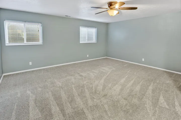Spacious room featuring light gray carpeting, two windows with horizontal blinds, and a bronze-finish ceiling fan with integrated lighting