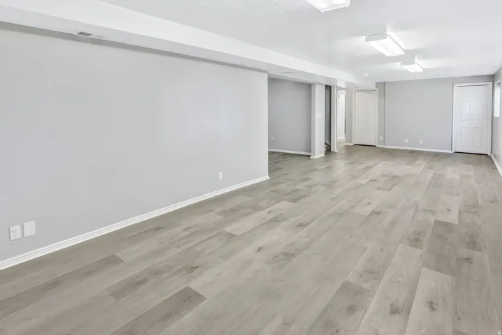Spacious room featuring light wood-finish flooring, neutral gray walls, white baseboards, and overhead fluorescent lighting
