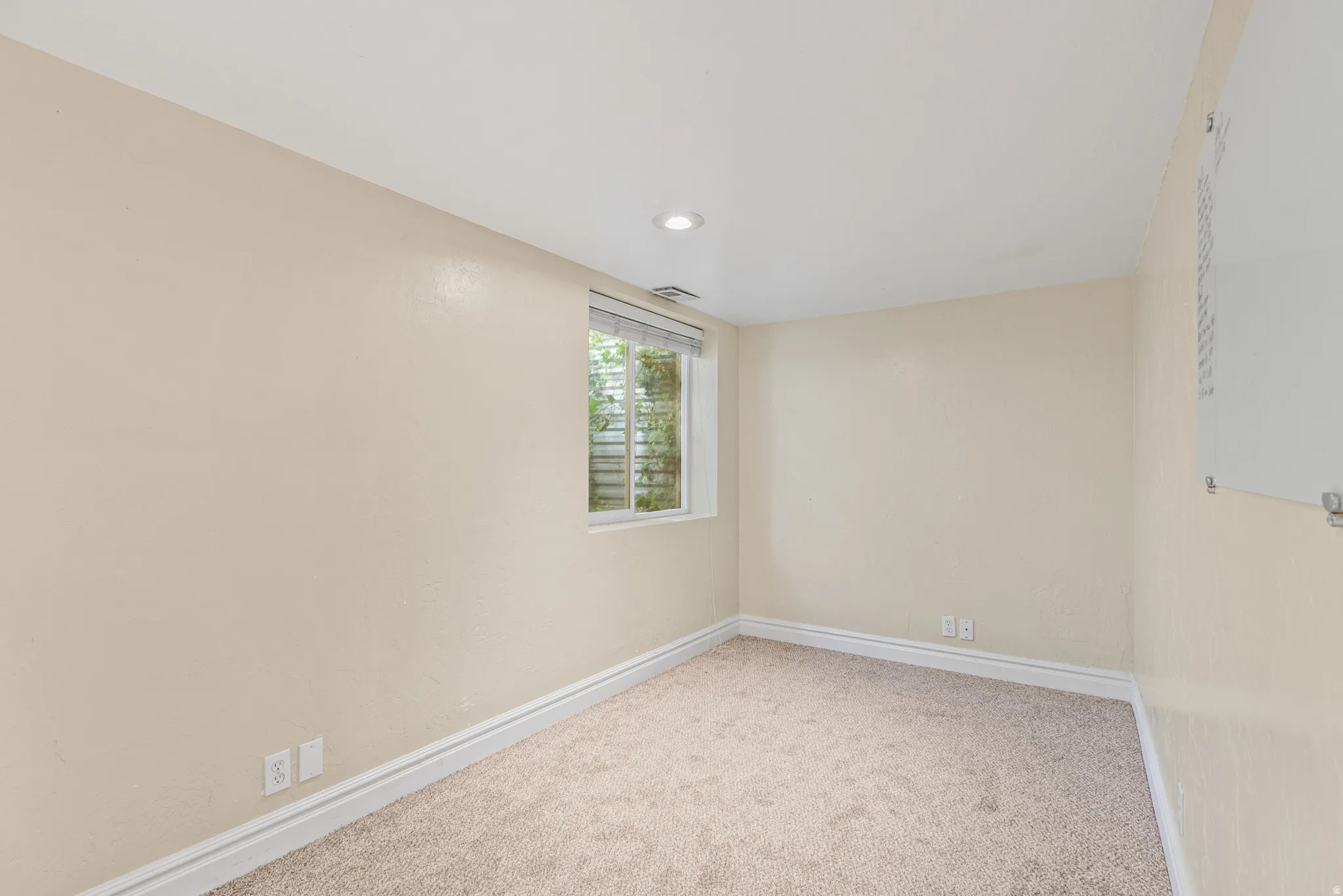 Recessed ceiling lighting, a window with exterior foliage, neutral-toned walls, and plush carpeting