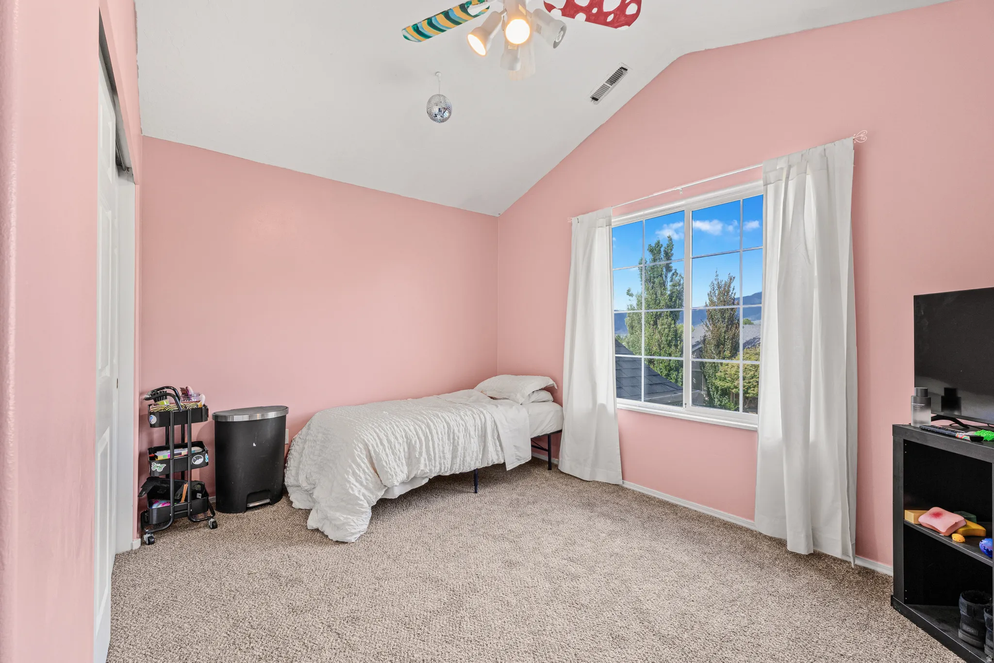 Vaulted ceiling, large window with multi-pane grid, neutral carpet flooring, and a ceiling fan