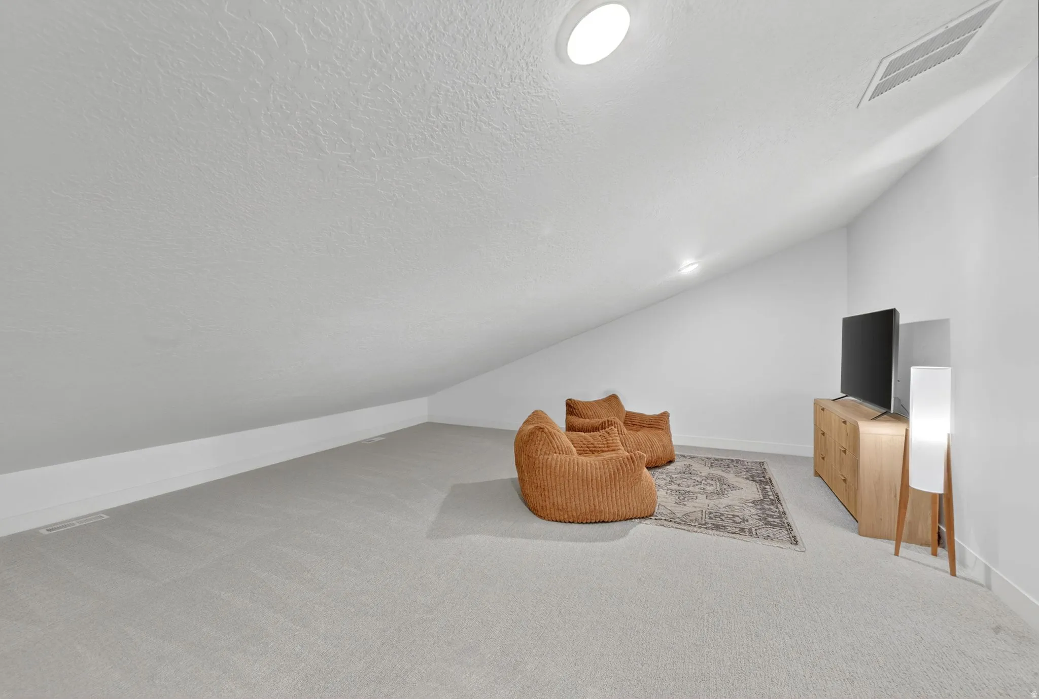 Spacious loft area featuring light gray carpeting, vaulted ceilings, a built-in recessed light fixture, and a wall-mounted air vent