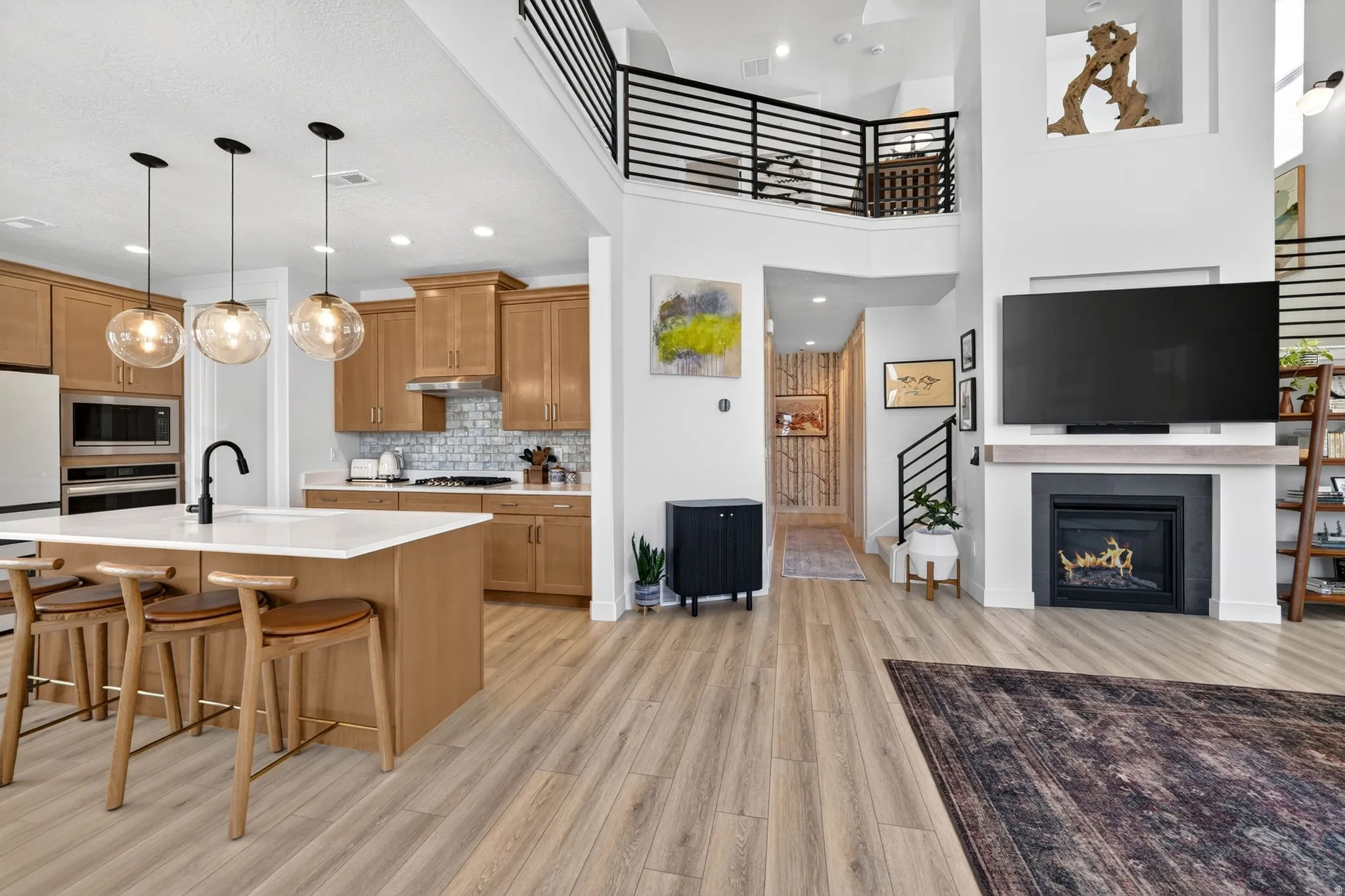Open-concept living space featuring wood-finish flooring, a contemporary kitchen with light wood cabinetry and a central island, and a modern fireplace with a mantel