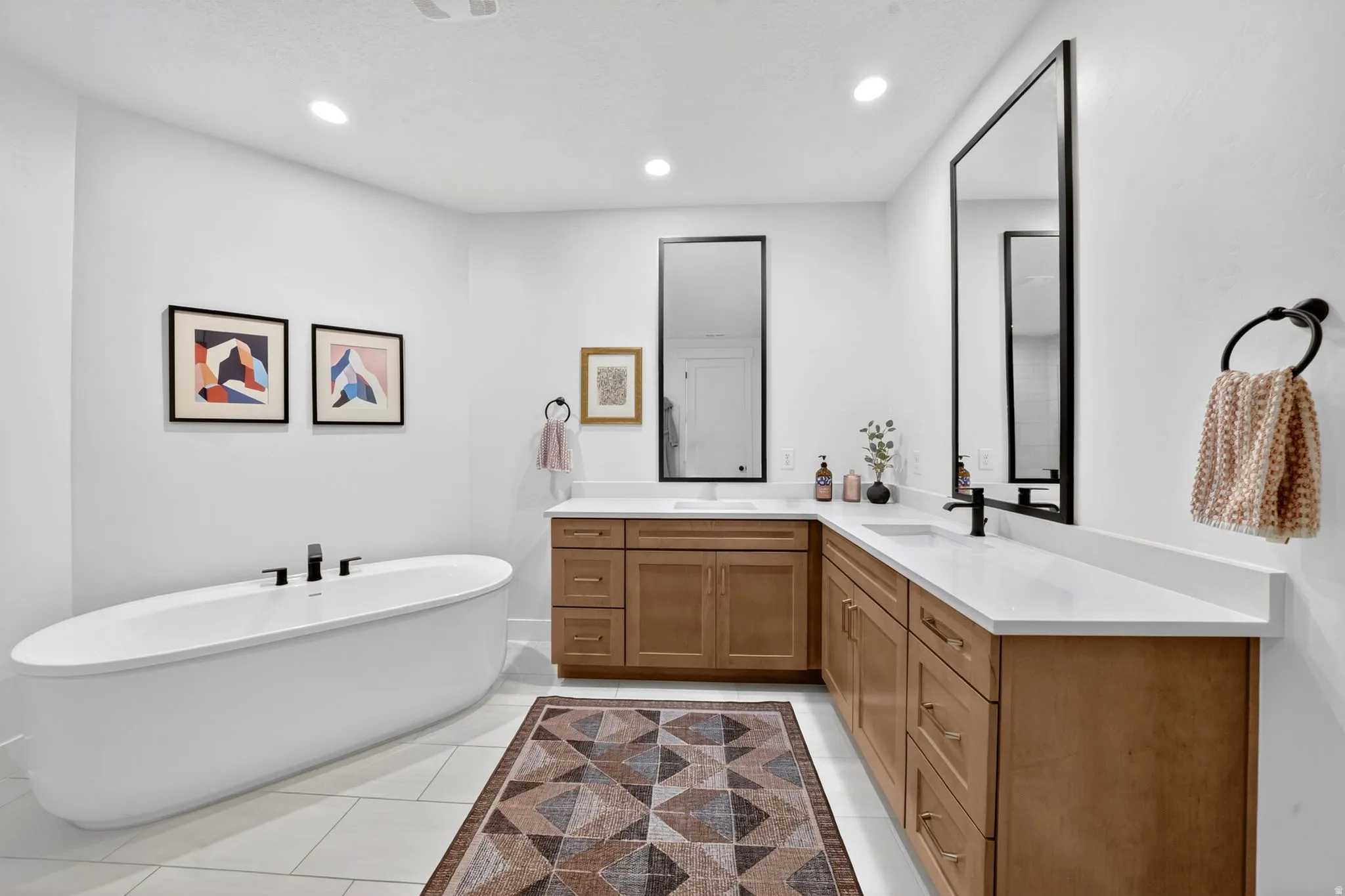 Spacious bathroom featuring a freestanding oval tub with matte black fixtures, an extensive wood-finish double vanity with white countertops and integrated sinks, large rectangular mirrors, and recessed lighting