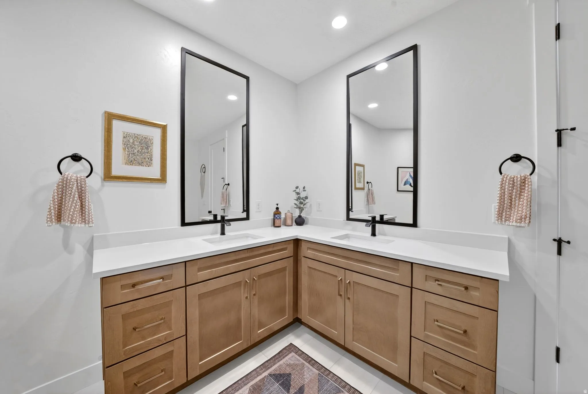 Corner vanity featuring dual sinks, white countertops, wood-finish cabinetry, black framed mirrors, and matte black fixtures