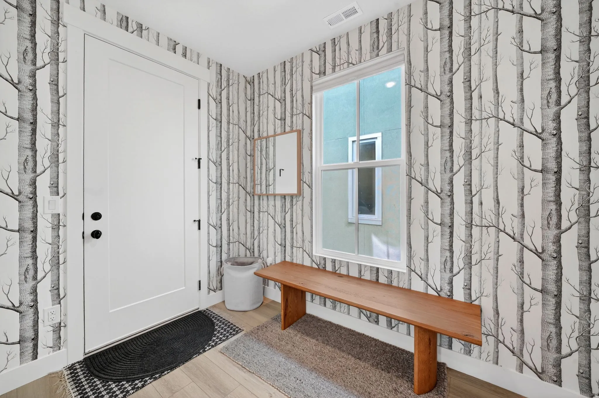 Entryway featuring tree-patterned wall covering, a white panel door with black hardware, and wood-finish flooring