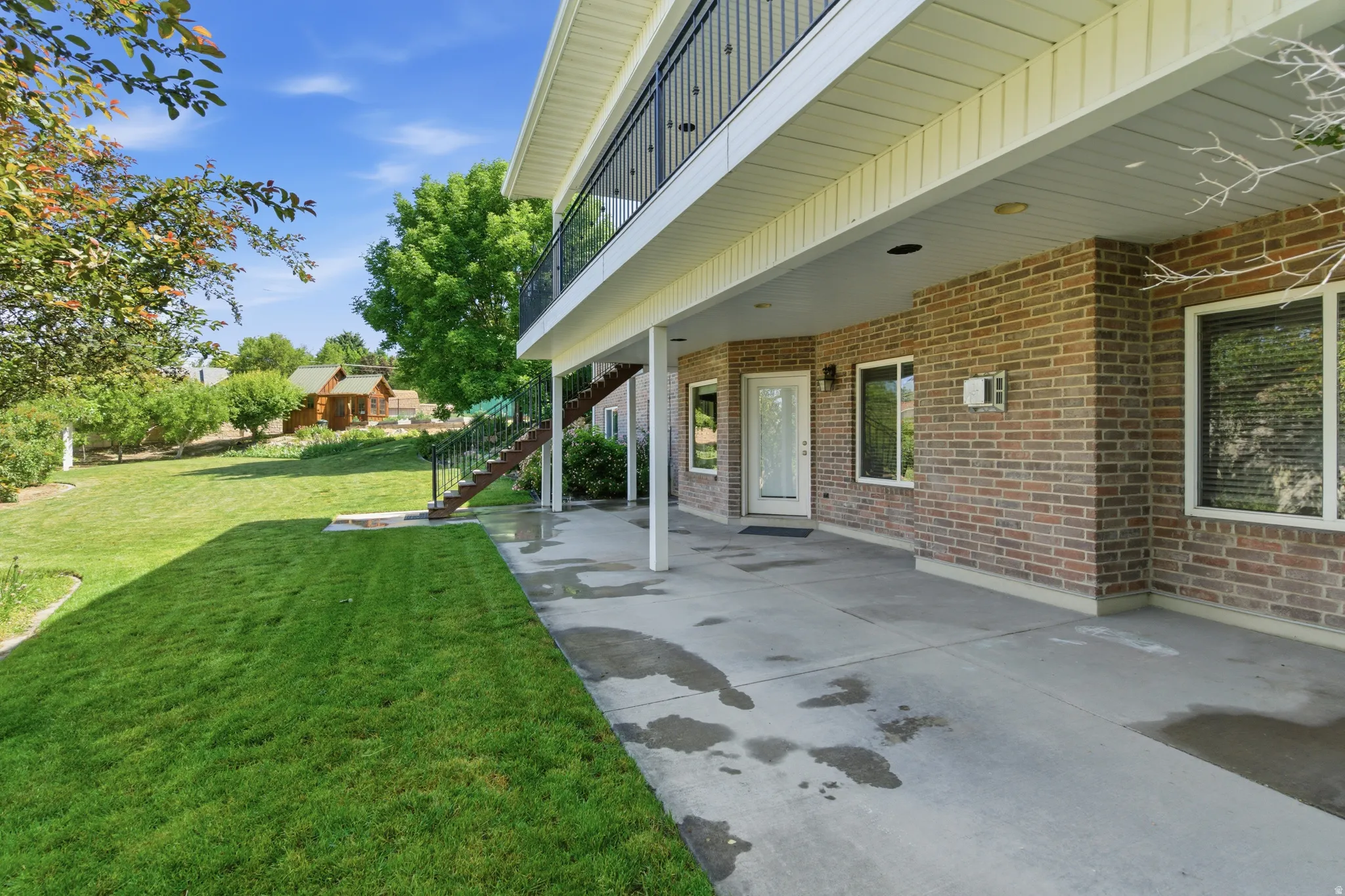 Expansive concrete patio with brick exterior and white trim, featuring a glass-paneled entry door, multiple windows, and a black wrought-iron balcony above
