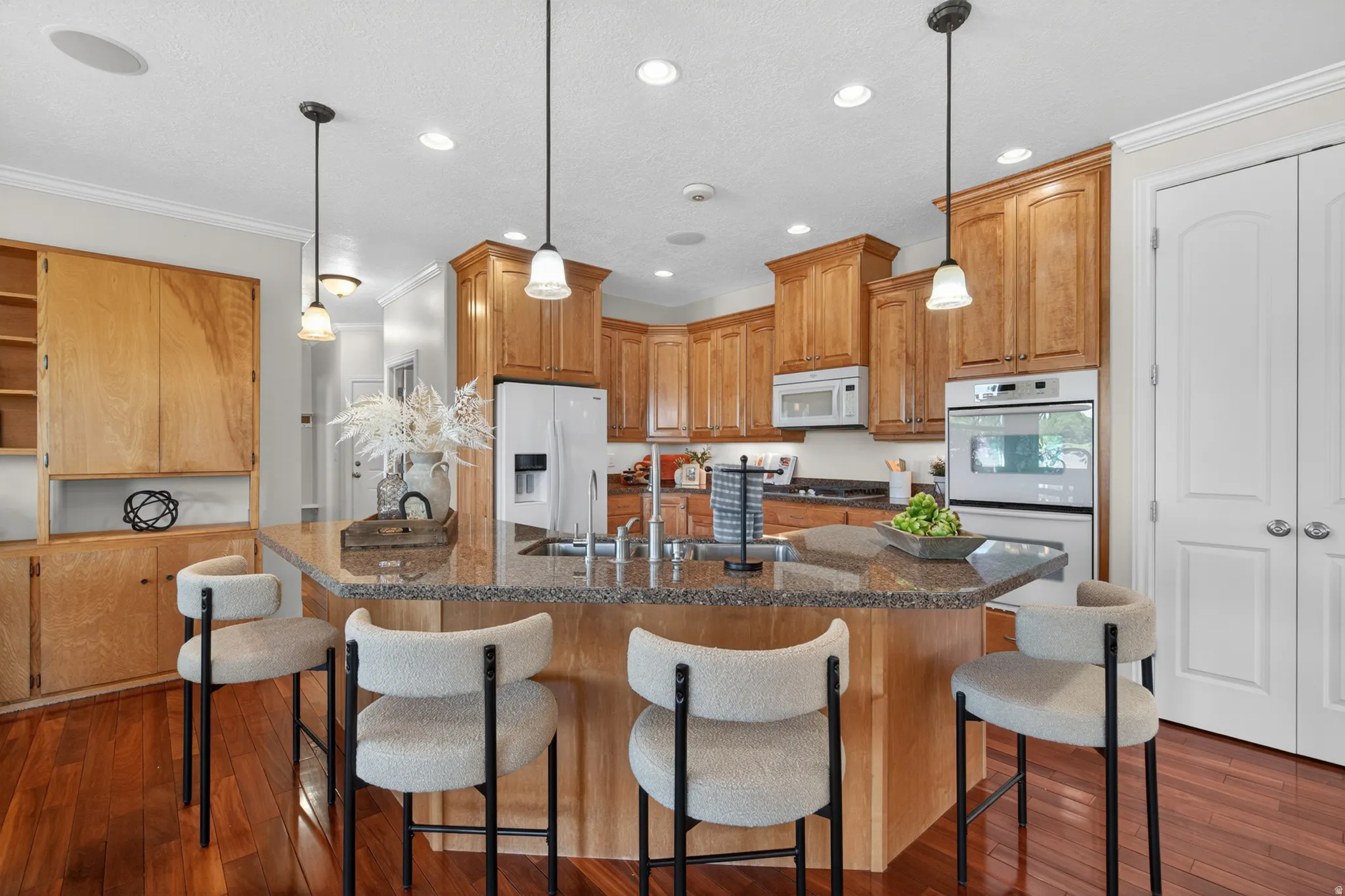 Kitchen featuring warm wood cabinetry, granite countertops, a central island with pendant lighting, integrated white appliances, and rich wood-finish flooring