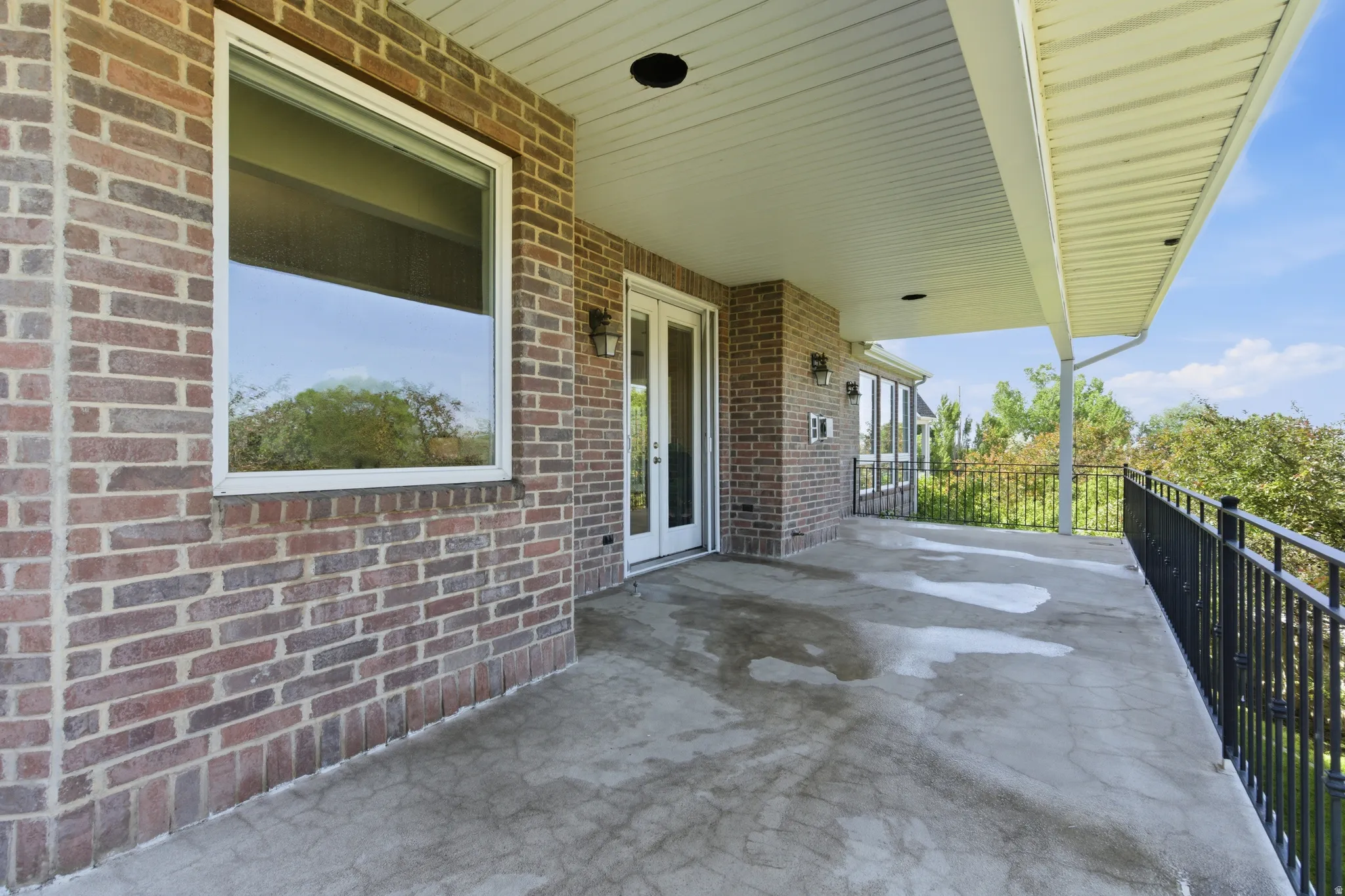 Expansive covered concrete patio featuring a brick exterior wall, white framed window, recessed lighting, and black metal railing