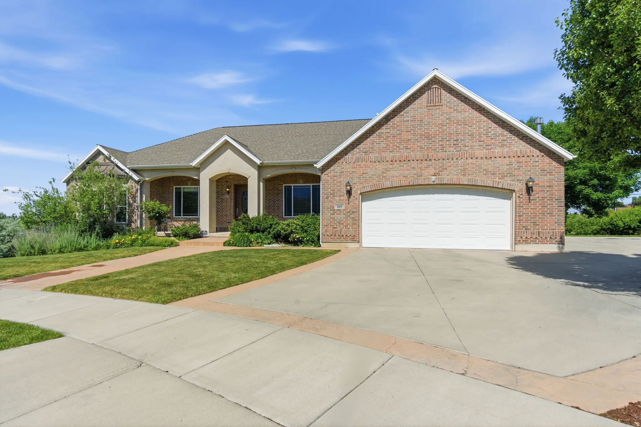 Brick and stucco exterior featuring a two-car garage, mature landscaping, a paved driveway, and a concrete pathway to the main entry