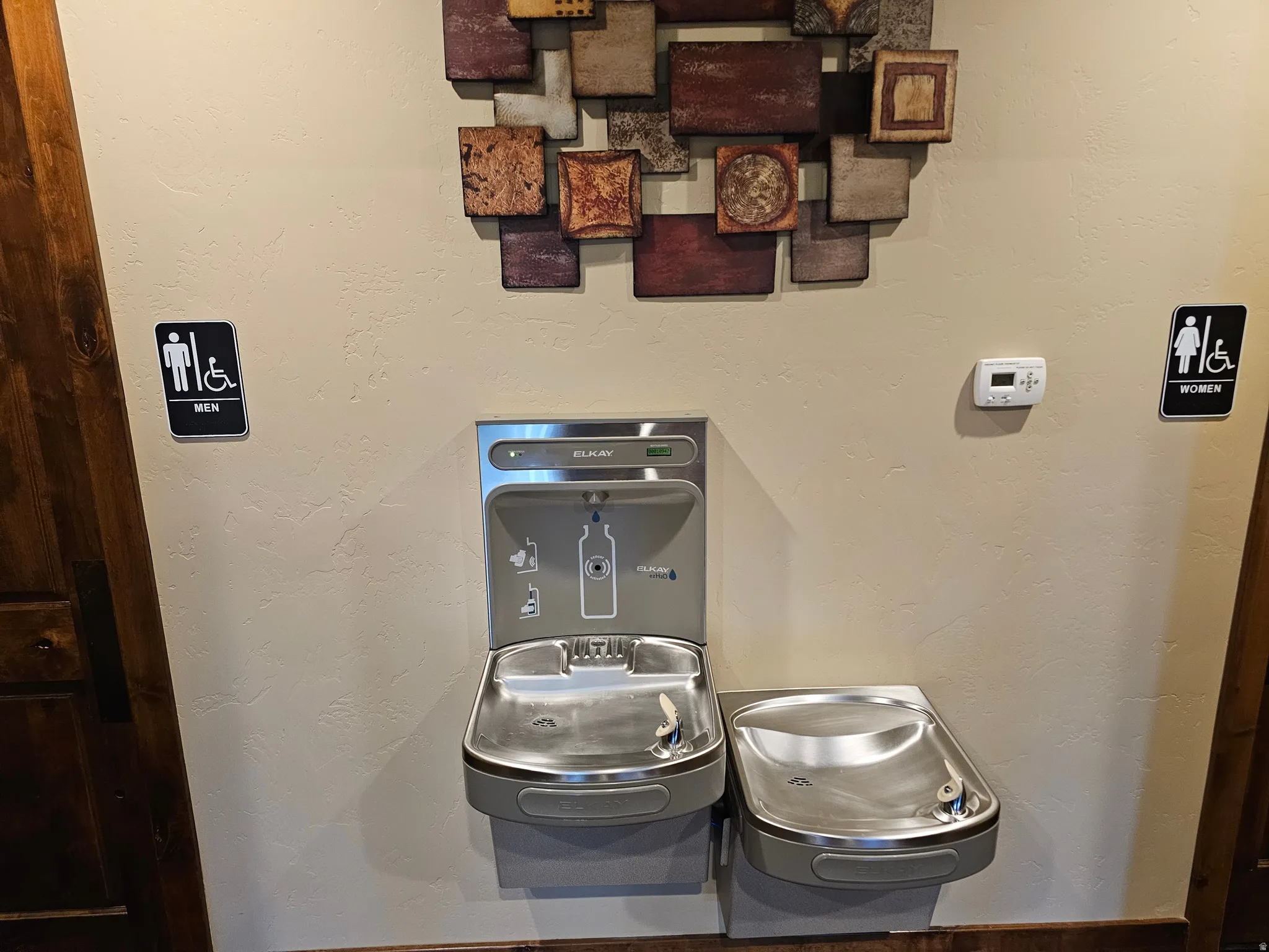 Dual stainless steel drinking fountain with bottle filler, set against a neutral-toned wall with a textured art installation