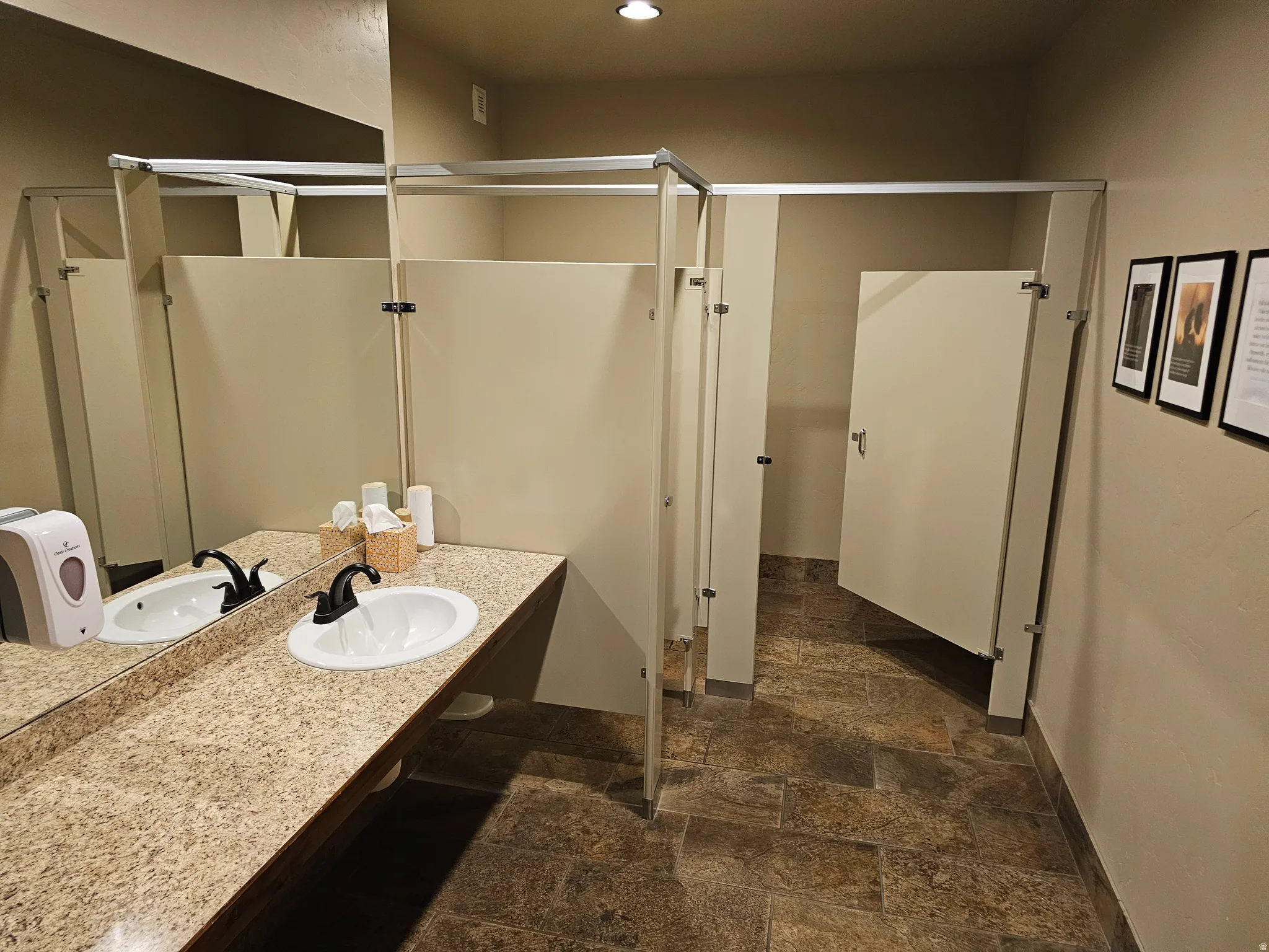 Spacious restroom featuring a stone-look tile floor, light-colored partition walls, and a long vanity with two sinks