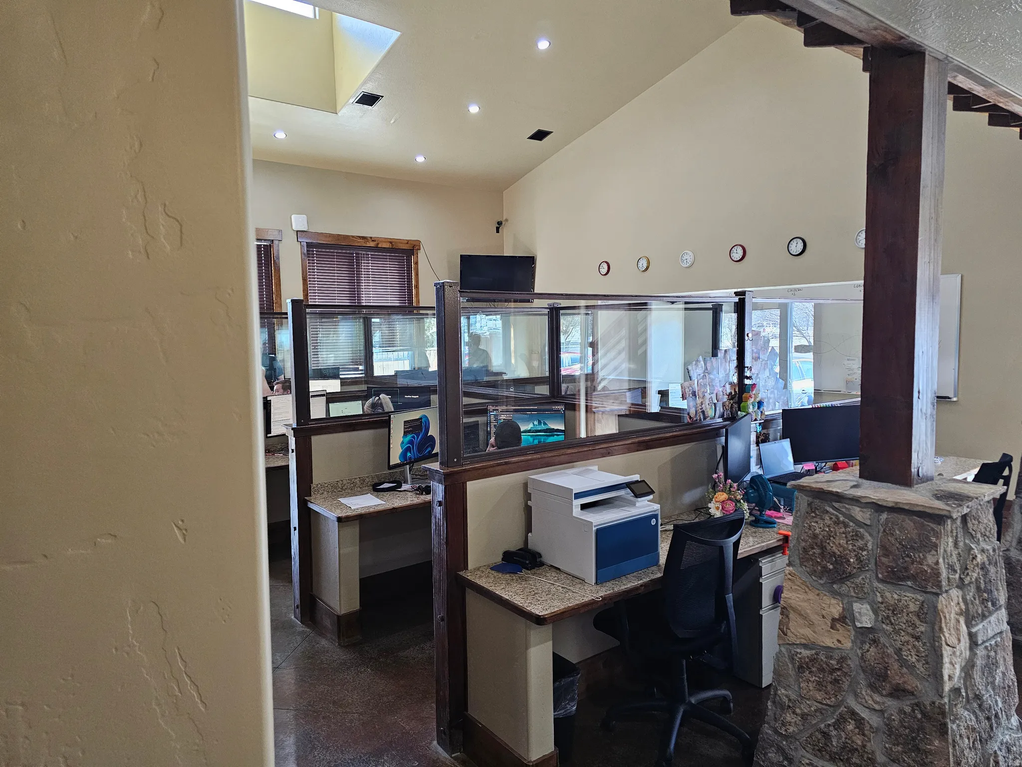 Open office space featuring cubicle workstations with granite-finish countertops and glass partitions, vaulted ceilings, recessed lighting, and a prominent stone-clad column